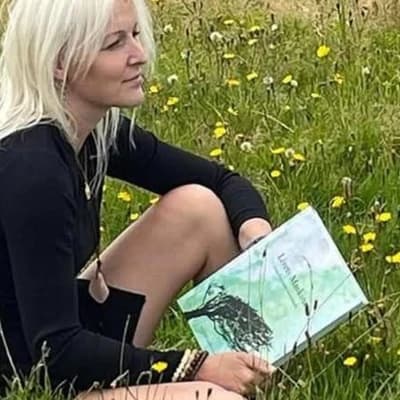 Lisa S. Andersen sidder i en mark med mælkebøtter og holder Livets Maskine i hånden
